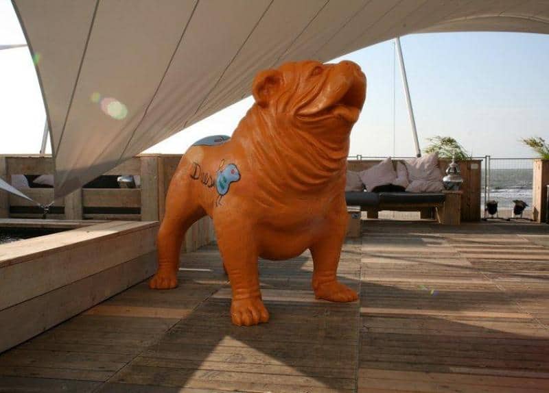 Große Bulldoggenfigur 1 Pomarańczowa statua psa na drewnianym tarasie pod białym zadaszeniem.