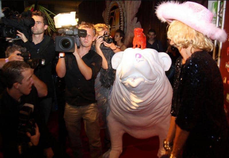 Große Bulldoggenfigur 2 Kobieta w różowym kapeluszu pozuje na czerwonym dywanie obok dużej, białej rzeźby psa, otoczona przez fotoreporterów.