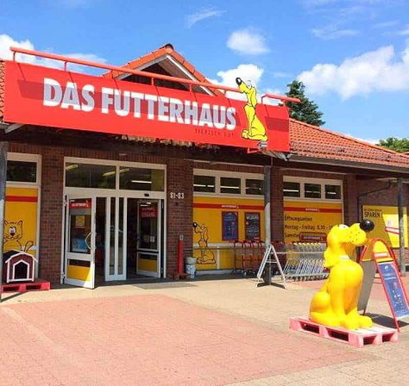 Werbehund FUTTERHAUS 2 Sklep Das Futterhaus z czerwonym szyldem nad wejściem, żółtą figurką psa i plakatami.