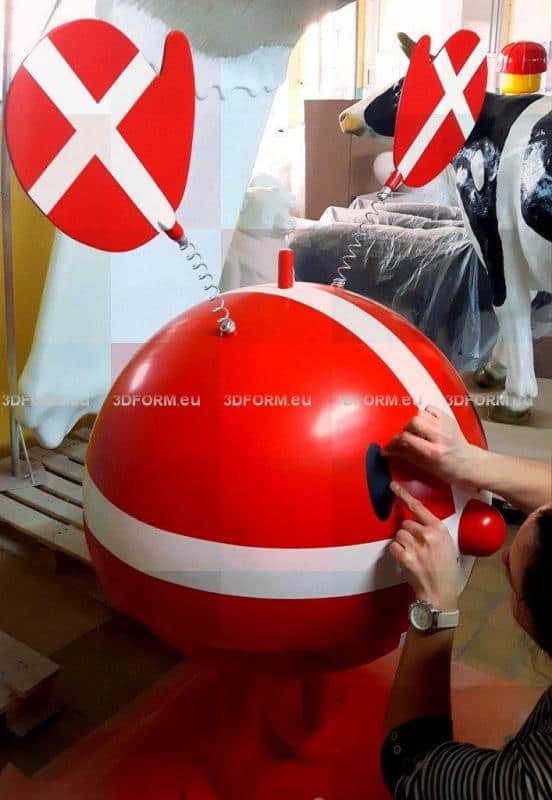 Werbefigur Hoptimist 1 Czerwona, okrągła figura z uszami na sprężynach i białymi paskami, z rękami przymocowującymi oko. W tle plastikowa krowa.