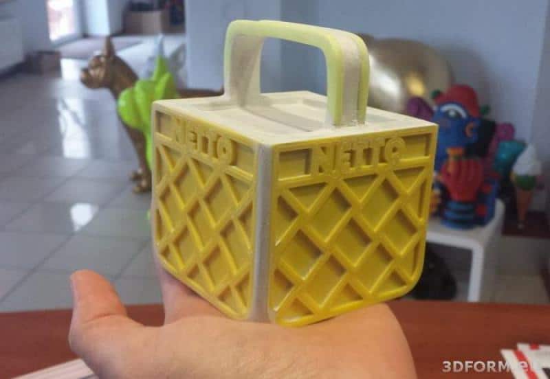 NETTO 3D-LOGO 4 Mały model żółtej skrzynki z napisem "NETTO", trzymany na dłoni.