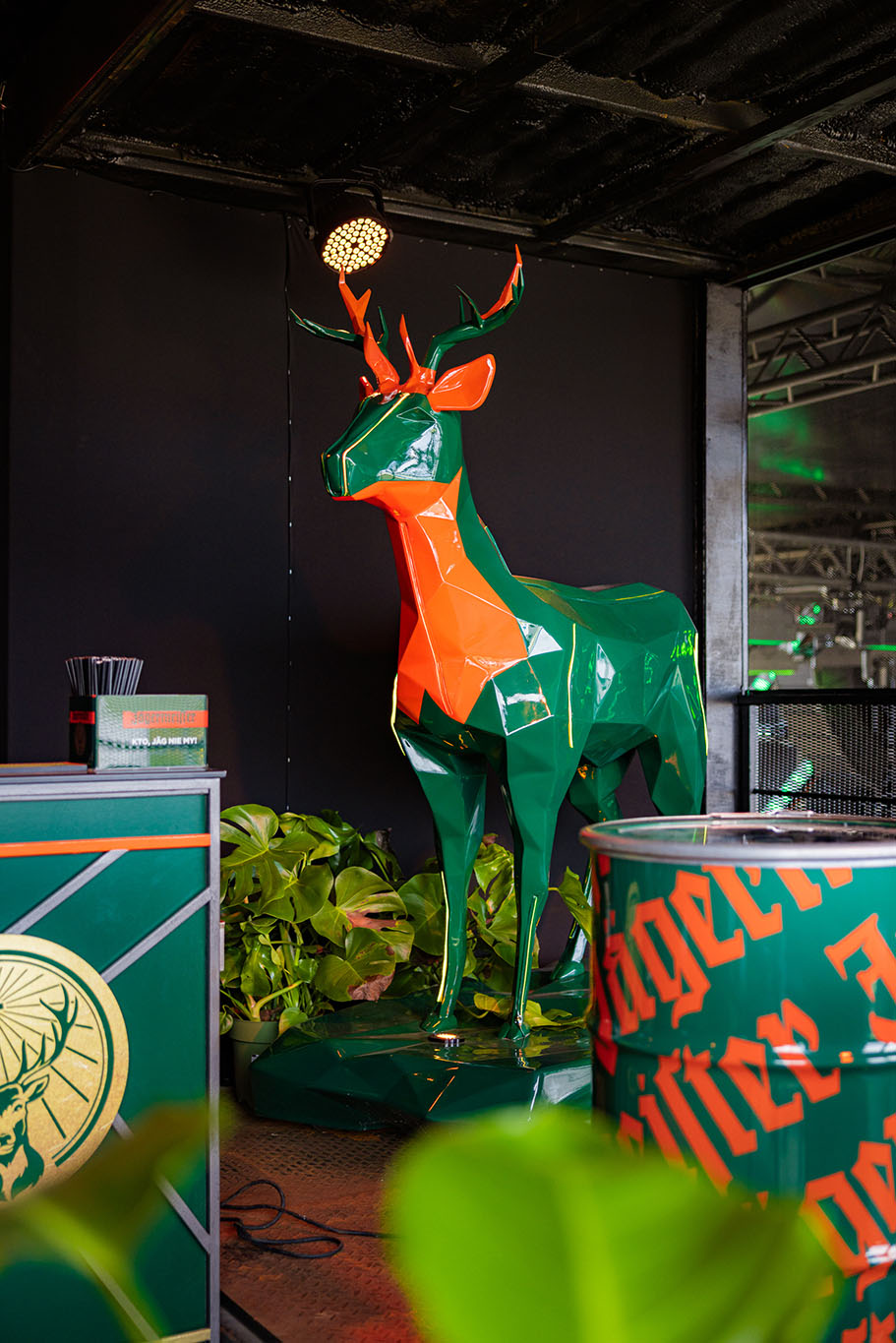 Jägermeister-Hirsche mit LED-Beleuchtung 2 Pomarańczowo-zielona rzeźba jelenia stojąca w otoczeniu roślin i zielono-pomarańczowych elementów dekoracyjnych.