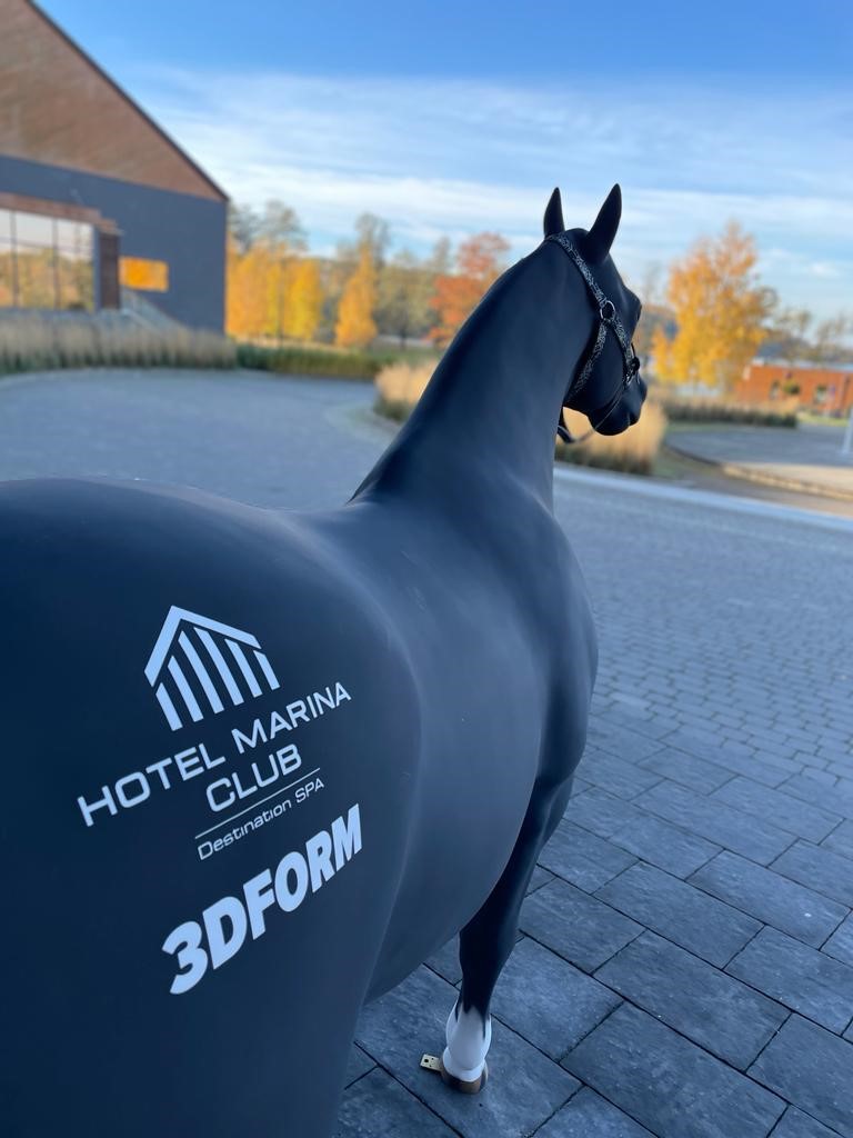 Pferd für das Marina Club Hotel 1 Figura czarnego konia z białym napisem HOTEL MARINA CLUB Destination SPA 3DFORM na grzbiecie, stojąca na bruku na tle jesiennego krajobrazu z budynkami i drzewami o żółtych liściach