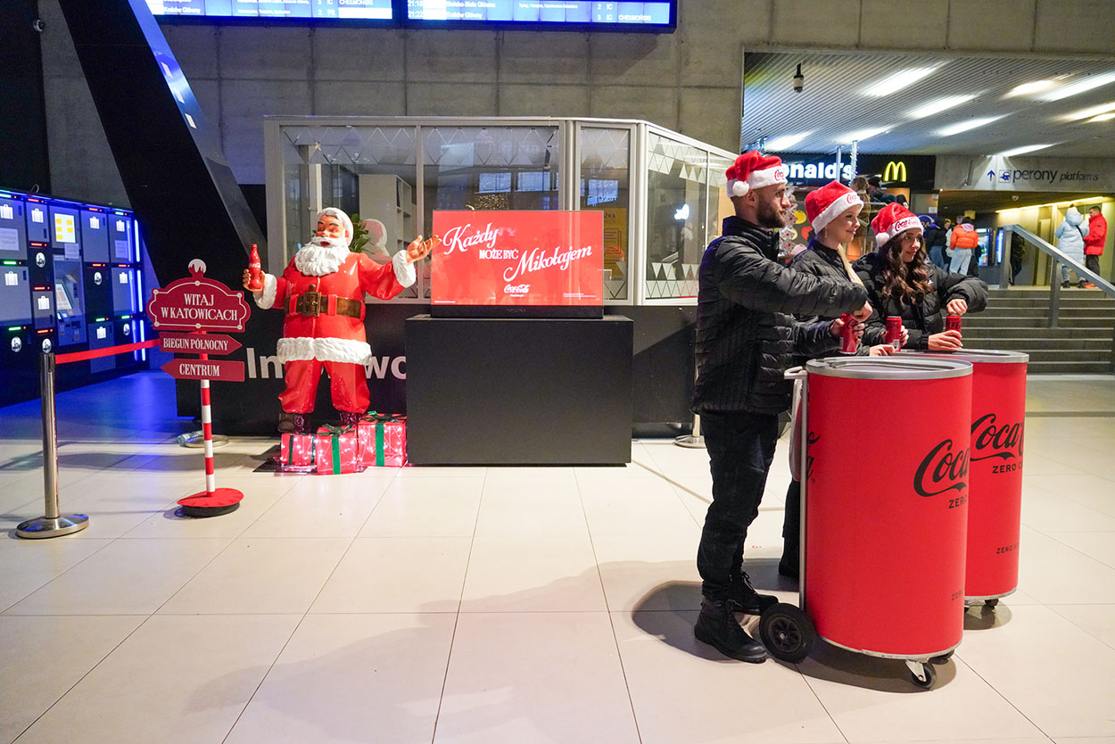 Jeder kann der Weihnachtsmann sein: Coca-Colas Weihnachtskampagne mit Weihnachtsmannfiguren 3 Trzy osoby w czapkach św. Mikołaja stoją przy czerwonych pojemnikach Coca-Coli w hali dworca, obok stoi figura św. Mikołaja z butelką Coca-Coli i znakiem "Witaj w Katowicach" oraz cyfrowym ekranem z napisem reklamowym.