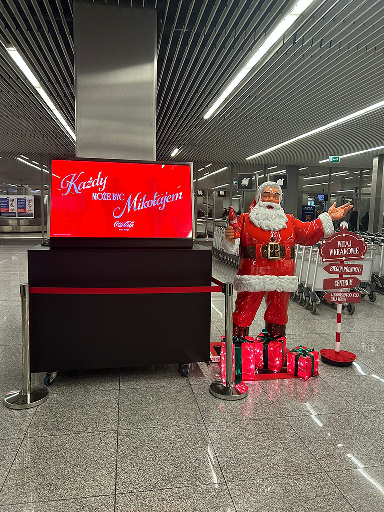 Jeder kann der Weihnachtsmann sein: Coca-Colas Weihnachtskampagne mit Weihnachtsmannfiguren 9 Figurka Świętego Mikołaja z butelką Coca-Coli i dekoracyjnymi prezentami przy informacji lotniskowej, obok stoi czerwony ekran z napisem "Każdy może być Mikołajem" oraz tablica powitalna z napisem "Witaj w Krakowie" w nowoczesnym terminalu lotniska.