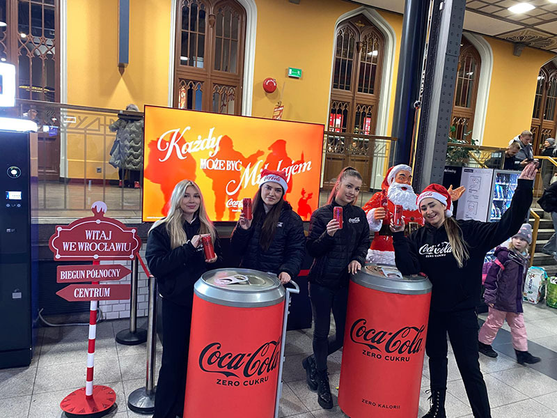 Jeder kann der Weihnachtsmann sein: Coca-Colas Weihnachtskampagne mit Weihnachtsmannfiguren 16 Cztery młode kobiety w ciemnych kurtkach i czapkach Mikołaja pozują z puszkami Coca-Coli przy dwóch dużych stojakach w kształcie puszek Coca-Coli Zero Cukru, na tle ekranu z napisem "Każdy może być Mikołajem" i figury Świętego Mikołaja.
