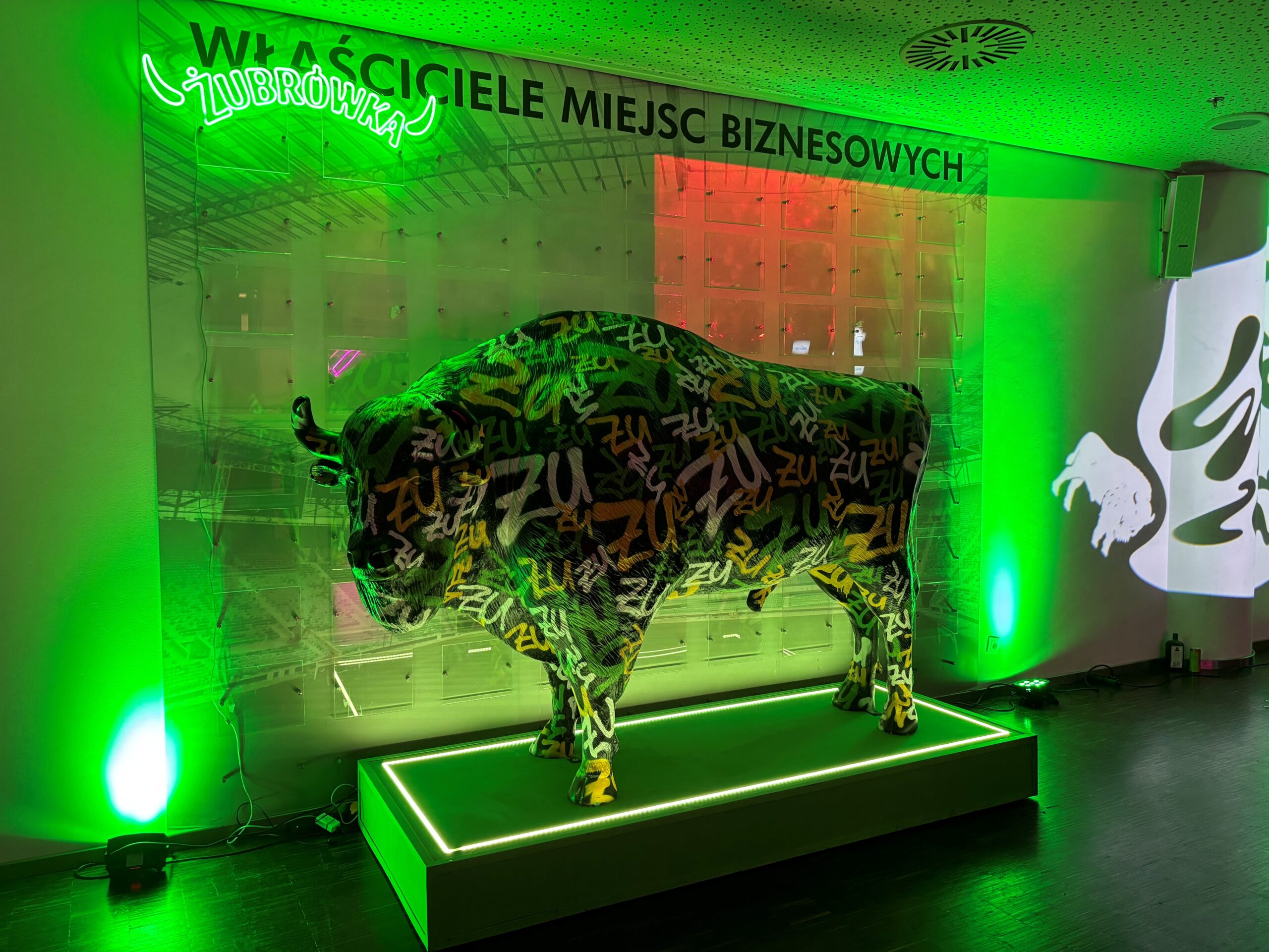 Wisent für die Wisent-Zone 1 Rzeźba bizona pokryta kolorowymi napisami „ZU” na podświetlanej zielonym światłem platformie, z neonowym napisem Żubrówka i napisem na ścianie "Właściciele miejsc biznesowych" w tle, w nowoczesnym wnętrzu z zielonym oświetleniem.